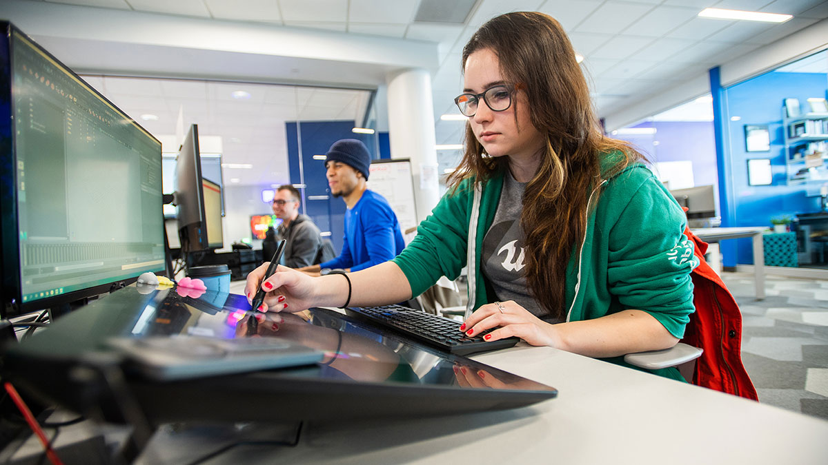 A young woman using a stylus to work on a design tablet in a modern computer lab.