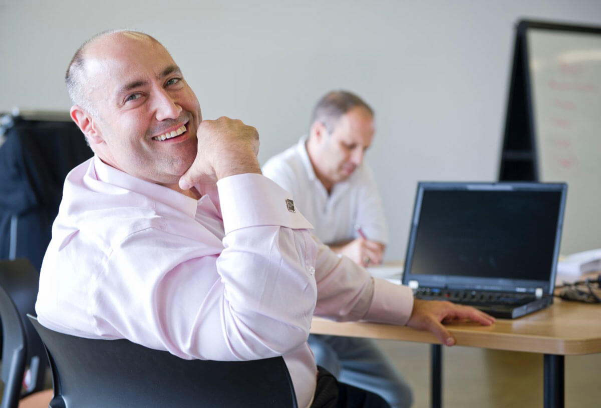 Two men sitting at a table, with one looking back toward the camera, smiling.