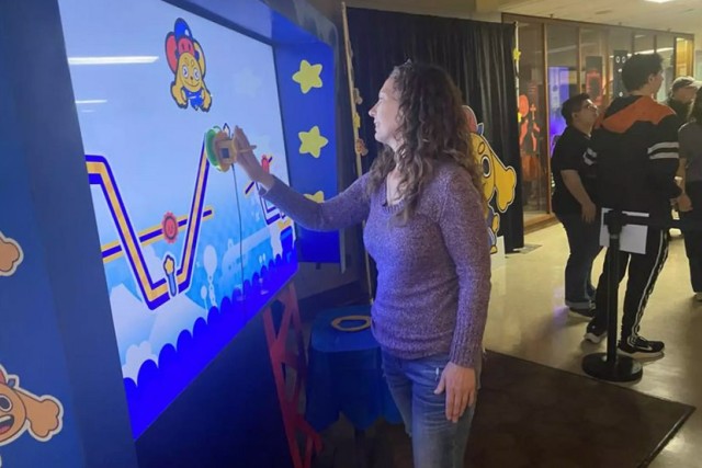 Carrie Principe playing the interactive Path Pursuit game on a large digital screen at Imagine RIT, with colorful cartoon graphics and other visitors watching nearby.