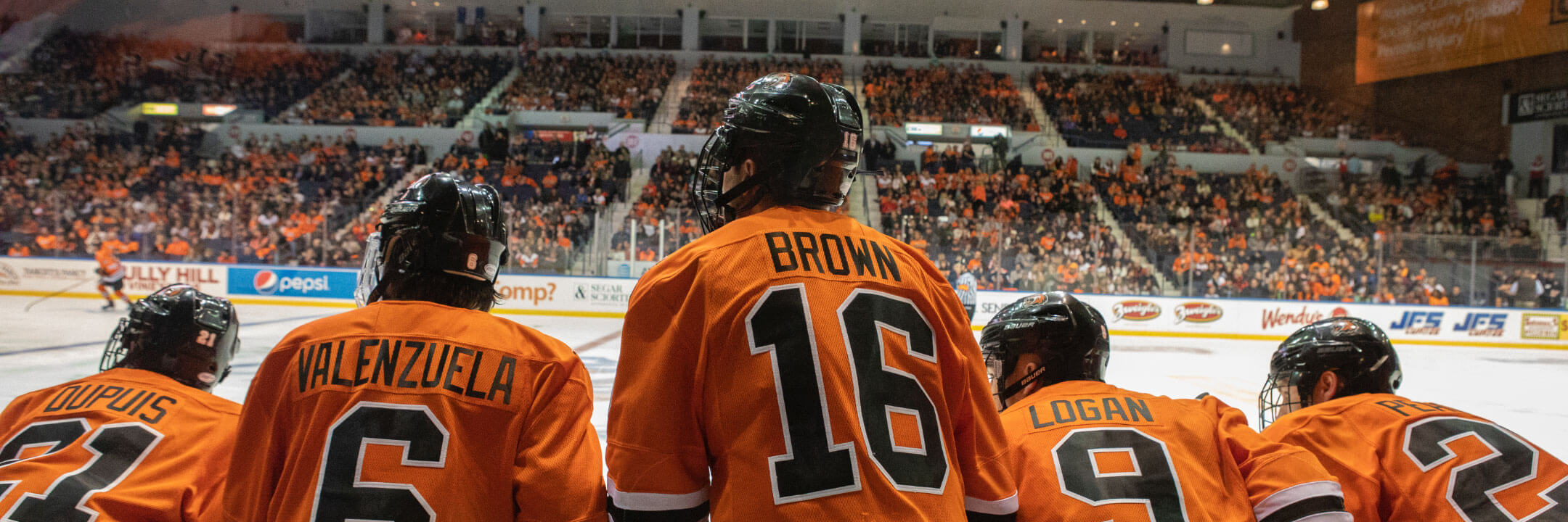 Five RIT hockey players looking out onto the ice with RIT fans in the crowd in the background..