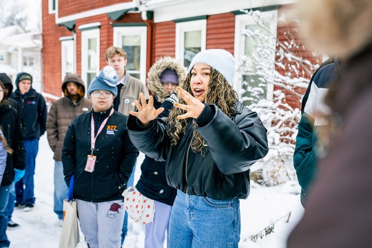 a college age female has her hands up in a circular fashion. She stands in a snowy area with other students and a red house behind her.