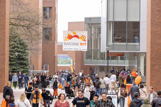 A large group of people walk on campus under an Imagine RIT banner.