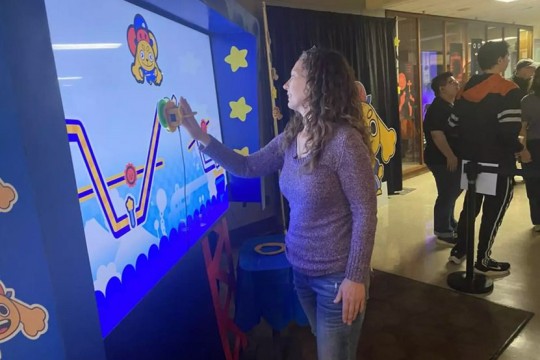 Carrie Principe playing the interactive Path Pursuit game on a large digital screen at Imagine RIT, with colorful cartoon graphics and other visitors watching nearby.