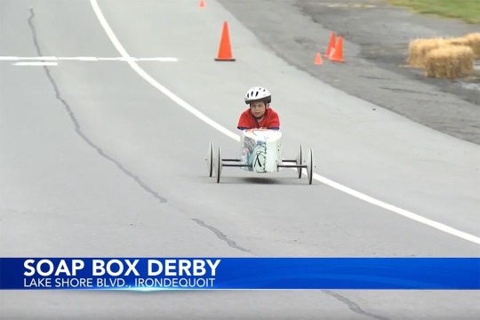 A screenshot from the TV news story shows a small child wearing a bike helmet as he rides down a hill in his Soap Box Derby car. Along the bottom of the screenshot is a news graphic that reads Soap Box Derby in big text, and Lake Shore BLVD., Irondequoit in smaller text directly below the large text.